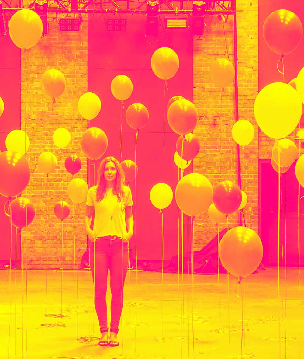A young woman standing among a dozen balloons.